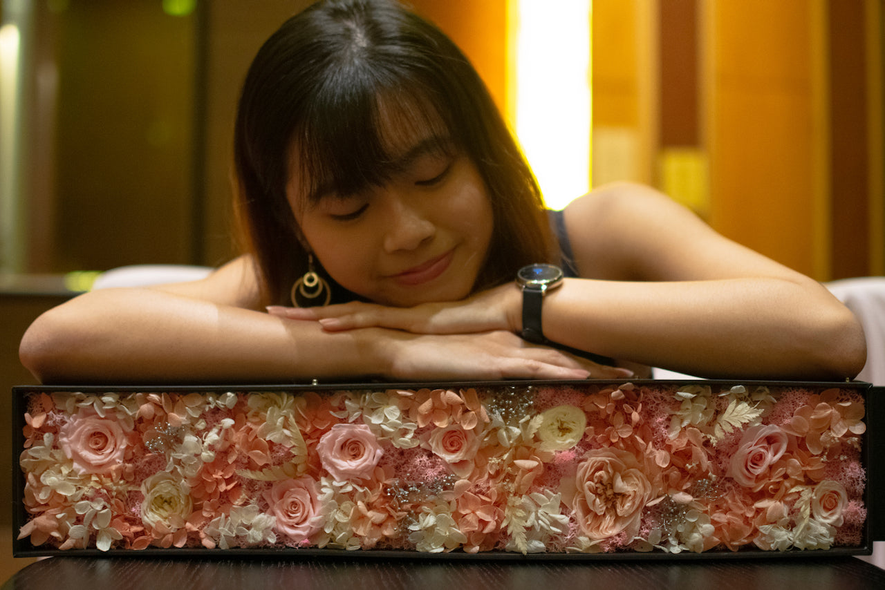 Young woman admiring a box of preserved pink and white flowers in a cozy indoor setting