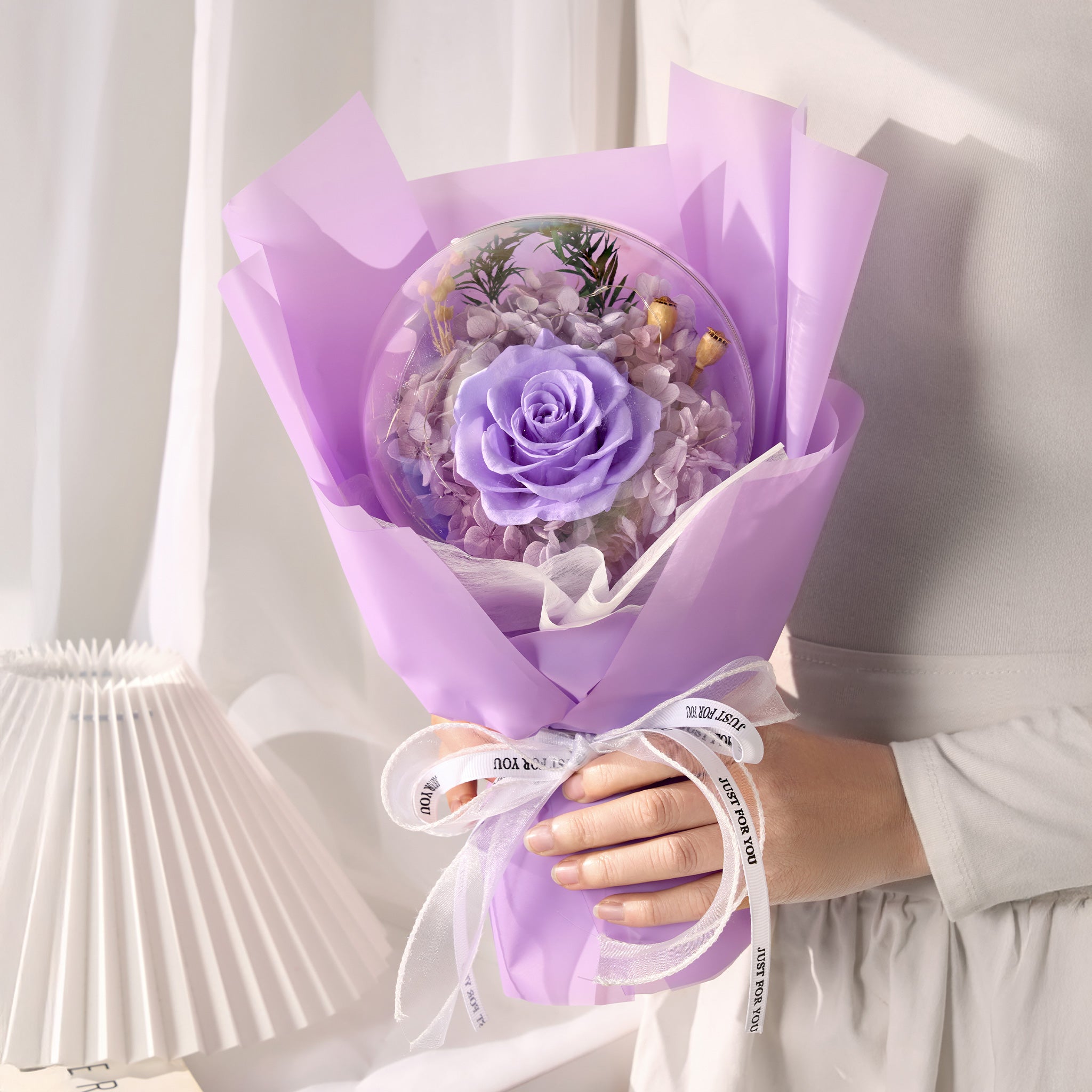 Bouquet of purple flowers encased in a transparent acrylic ball wrapped in purple paper held by a person.