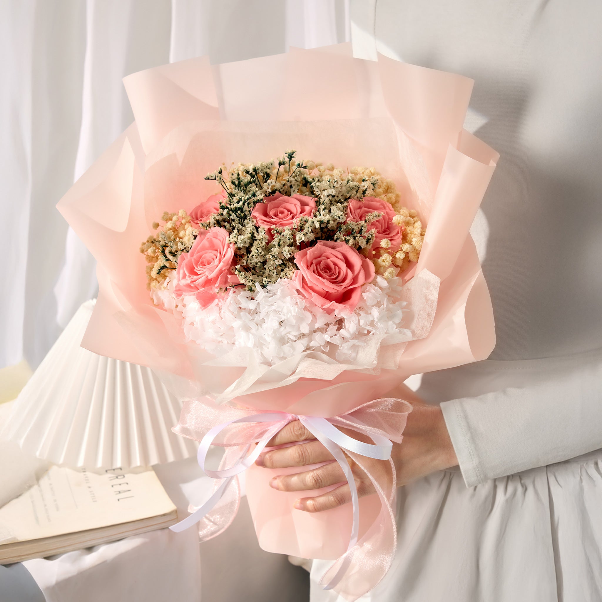 Bouquet of pink roses and greenery wrapped in light pink paper held by a person.