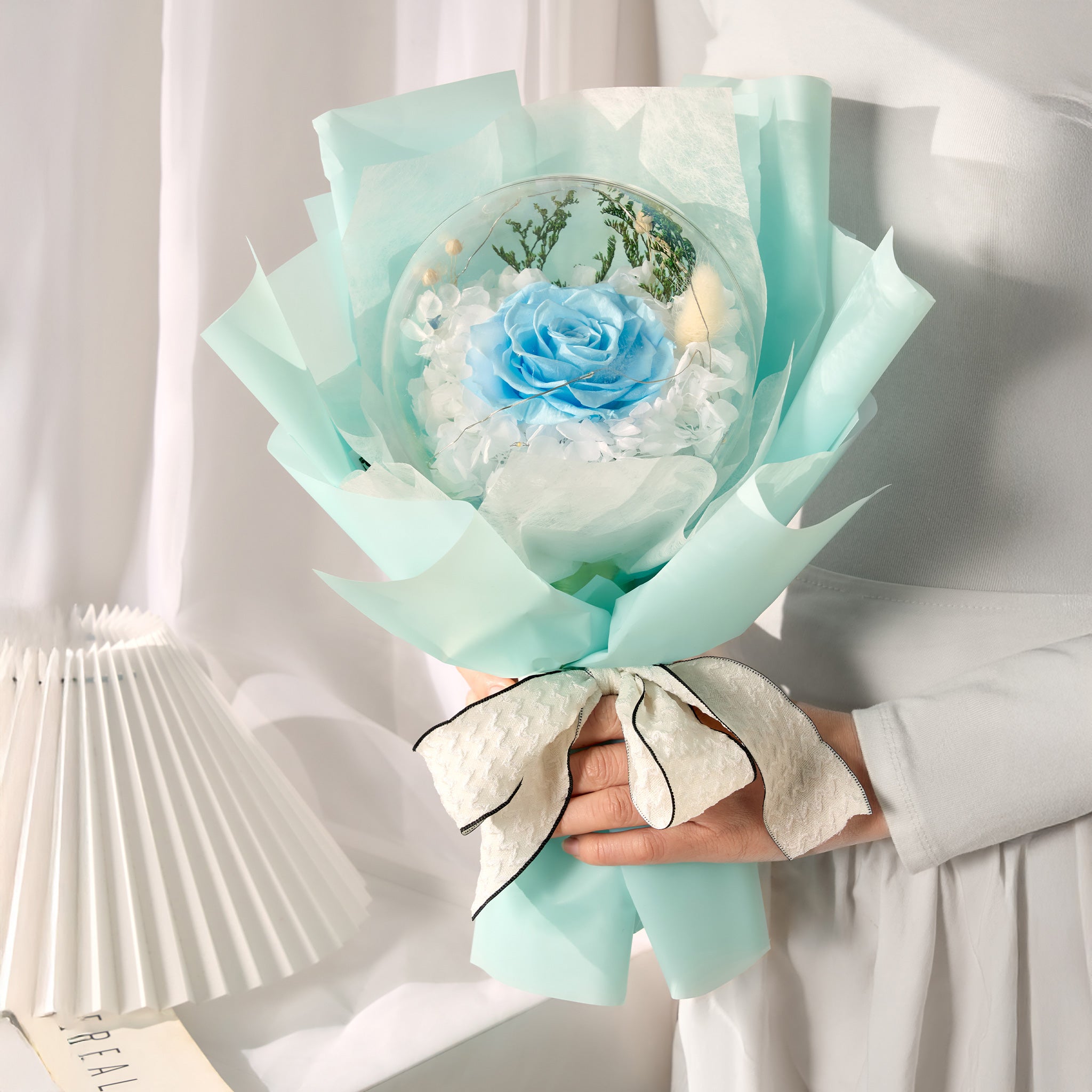 Bouquet of blue roses encased in an acrylic ball wrapped in light turquoise paper held by a person.